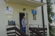 Kobieta w bluzce w paski wchodzi po schodach do budynku z numerem 7. Nad wejściem tablica informacyjna i kamera.