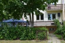 Dwupiętrowy budynek z balkonem, widoczny zza zielonych krzewów. Na balkonie biała balustrada. Z lewej strony widoczny fragment zadaszonego tarasu.