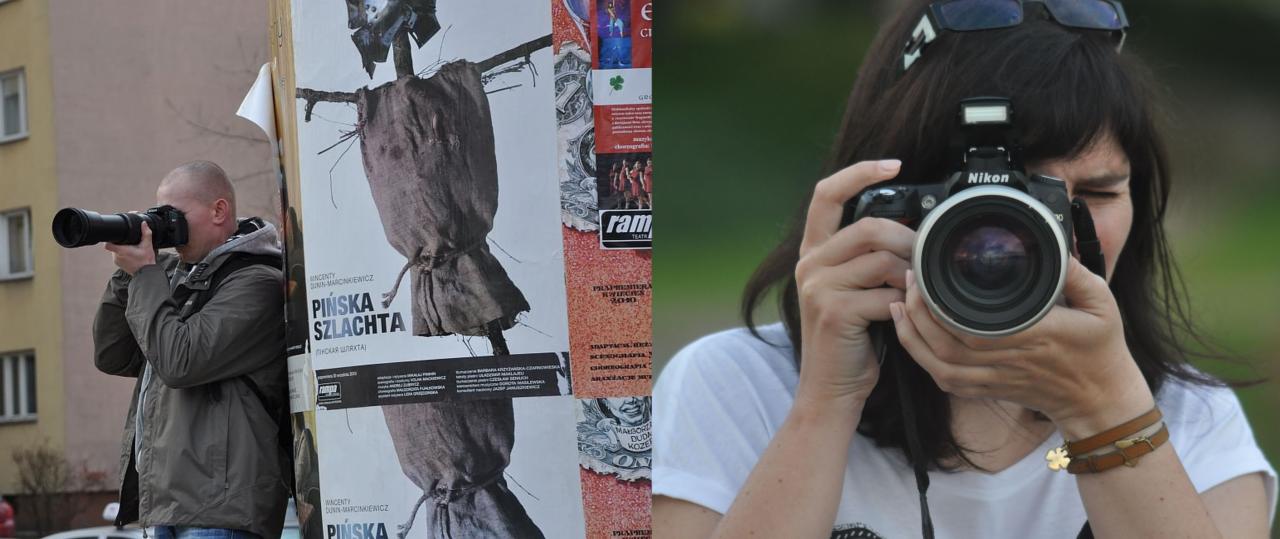 Dwie osoby fotografują. Mężczyzna z dużym obiektywem przy ścianie obklejonej plakatami, kobieta z aparatem Nikon, kadrująca przez obiektyw.