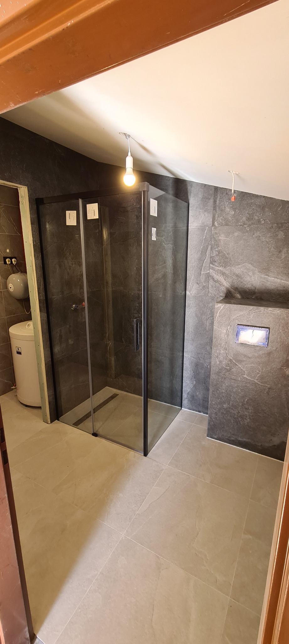 Nowoczesna kabina prysznicowa z czarnymi profilami i szarymi płytkami imitującymi kamień, z widocznym odpływem liniowym, zamontowana w łazience na poddaszu z białym bojlerem i wiszącą miską WC.