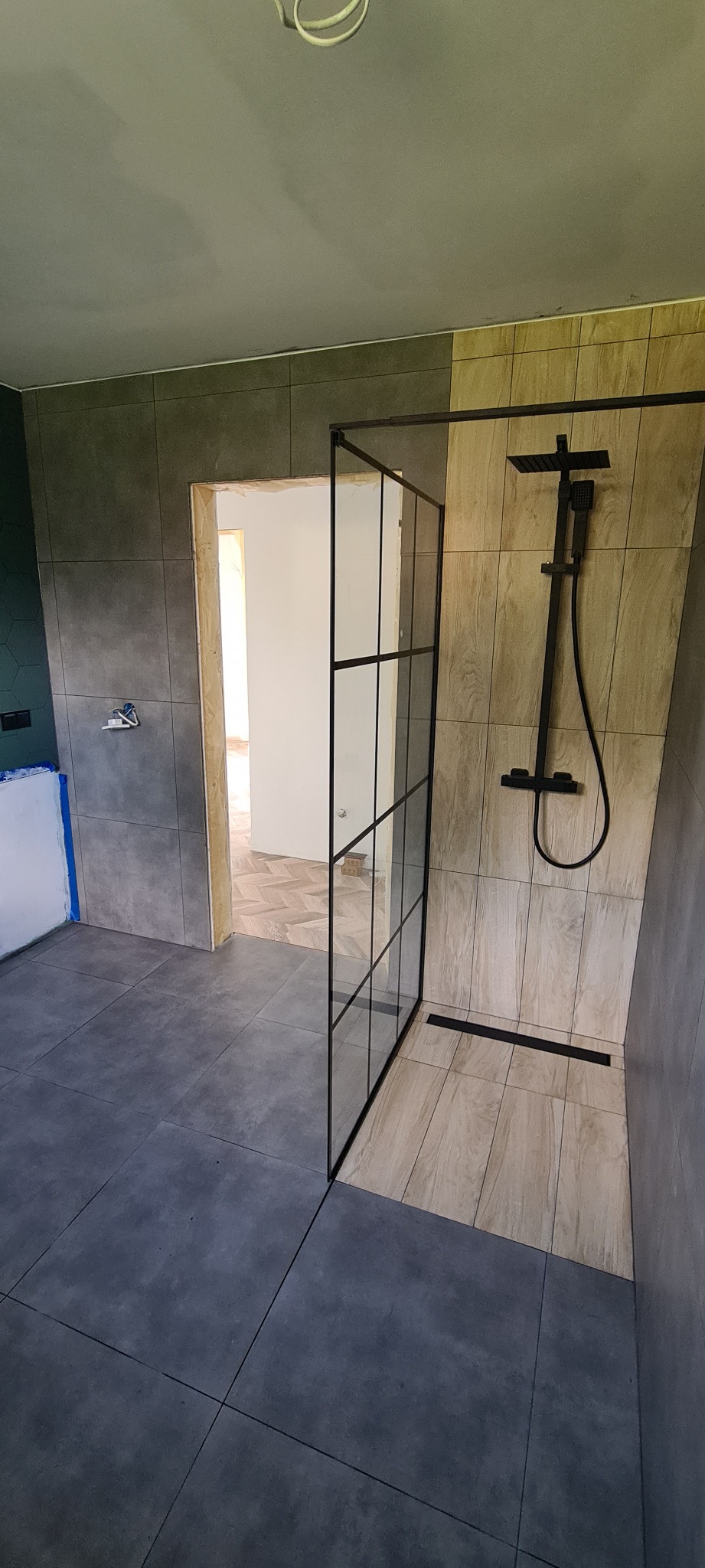 Nowoczesna łazienka w trakcie wykańczania z kabiną prysznicową typu walk-in z czarnymi profilami, widoczne szare i drewnopodobne płytki, oraz podłoga w jodełkę w sąsiednim pomieszczeniu.