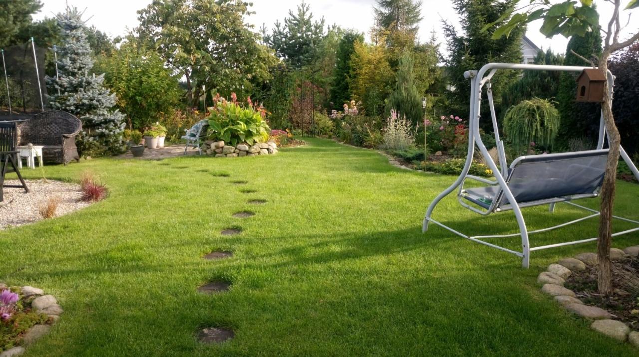 Zadbany trawnik z kamiennymi ścieżkami prowadzącymi do rabaty kwiatowej otoczonej kamieniami, w tle różnorodne drzewa i krzewy, po prawej stronie huśtawka ogrodowa z budką dla ptaków.