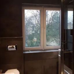 Wykończona łazienka z ciemnymi płytkami imitującymi kamień, jasną podłogą z płytek, białą stolarką okienną i kabiną prysznicową ze srebrnymi profilami.