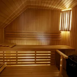 Wnętrze sauny wykończone jasnym drewnem, z ławkami na różnych poziomach, oświetleniem za oparciami i piecem do sauny w rogu.