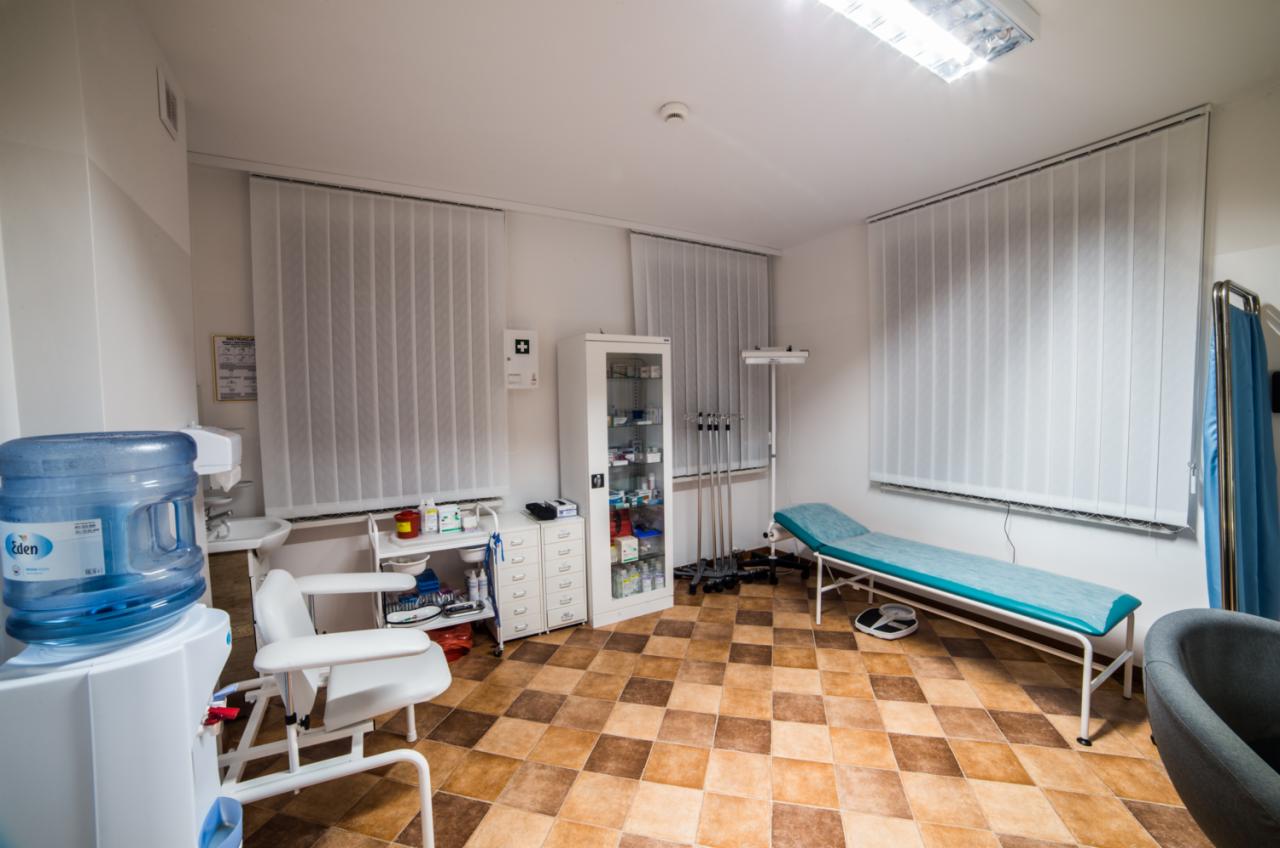 Jasne wnętrze gabinetu lekarskiego z leżanką, wagą, szafką z lekami i fotelem do pobierania krwi, widoczny dystrybutor wody Eden.