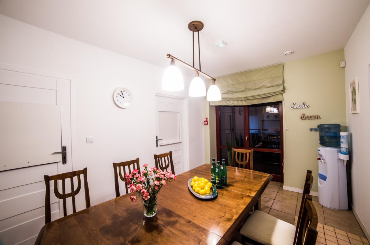 Jasne wnętrze pokoju z drewnianym stołem, bukietem różowych goździków w wazonie, butelkami wody i cytrynami na tacy, oświetlone wiszącą lampą z trzema kloszami, widoczne drzwi, zegar na ścianie...