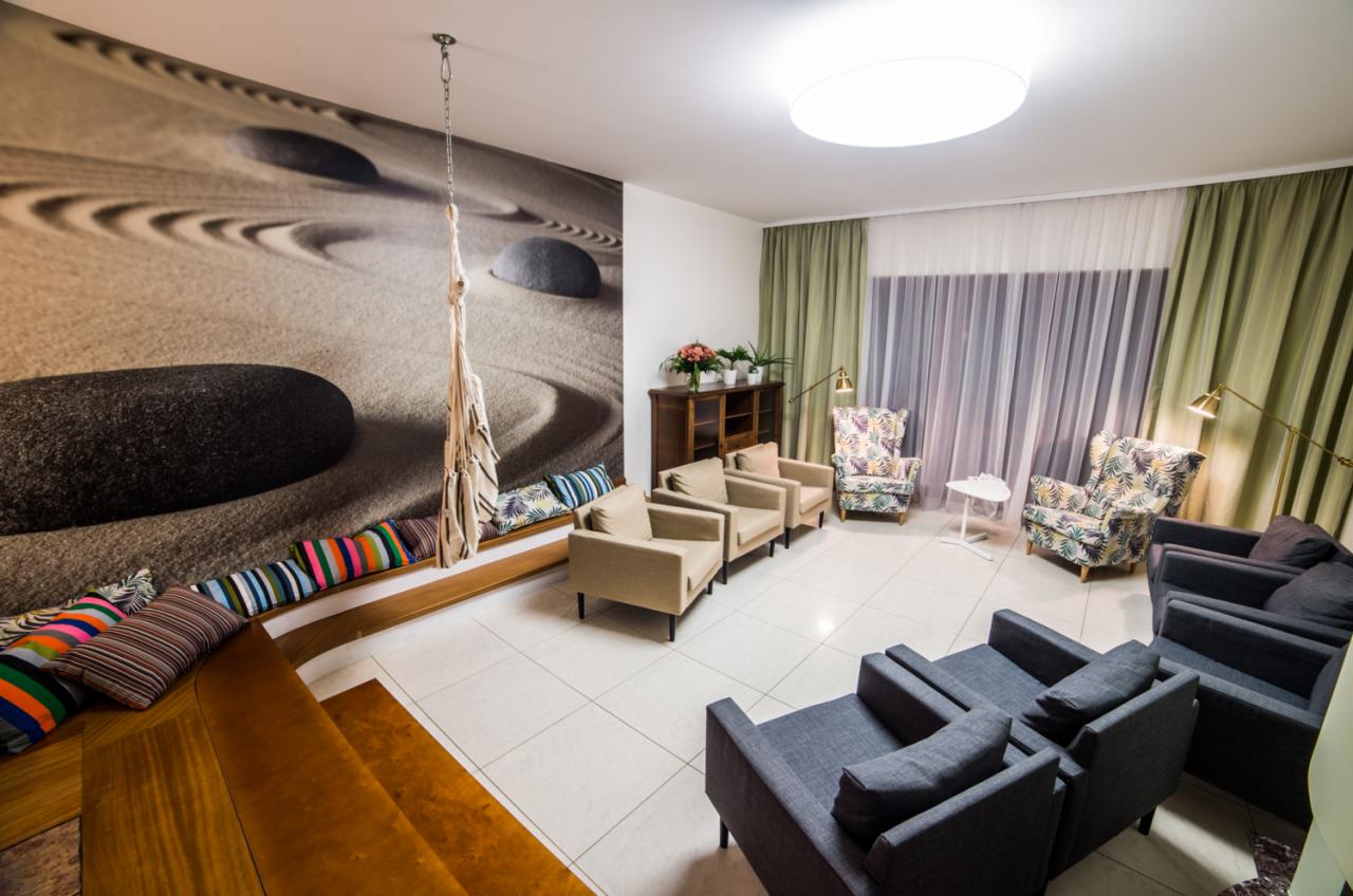 Jasne, przestronne wnętrze poczekalni z fototapetą zen, huśtawką hamakową, beżowymi i granatowymi fotelami oraz zielonymi zasłonami.