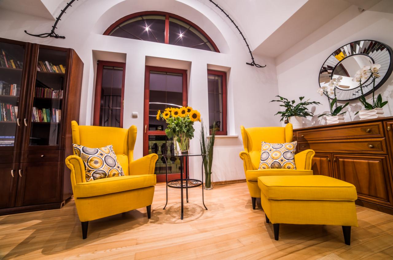 Jasne wnętrze z dwoma żółtymi fotelami typu uszak z podnóżkami, okrągłym lustrem na ścianie, bukietem słoneczników na szklanym stoliku i drewnianym regałem z książkami.