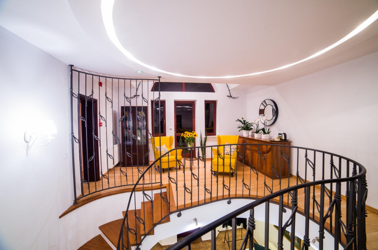 Wnętrze z drewnianymi schodami z kutą balustradą, żółtymi fotelami i dekoracyjnym oświetleniem sufitowym w jasnym pomieszczeniu.