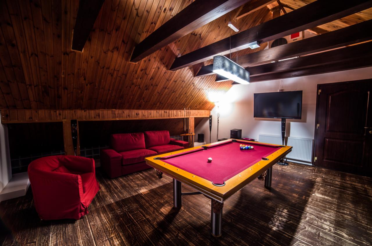 Pokój na poddaszu z drewnianą boazerią, belkami stropowymi, czerwonymi meblami wypoczynkowymi i stołem bilardowym z czerwonym suknem, oświetlony nowoczesną lampą.