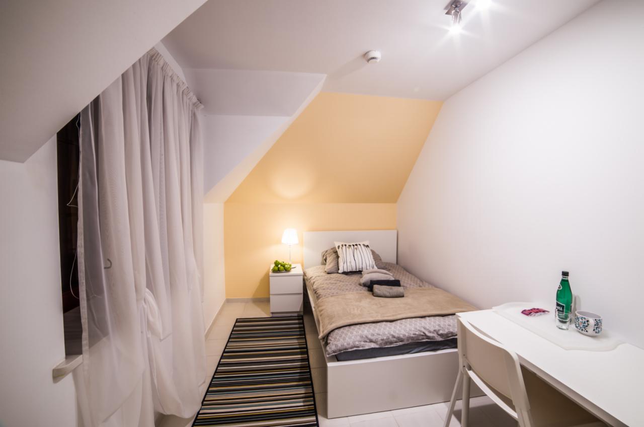 Pokój na poddaszu z łóżkiem pojedynczym, biurkiem, lampką nocną i zasłoniętym oknem, utrzymany w jasnej kolorystyce z akcentami pastelowej żółci.