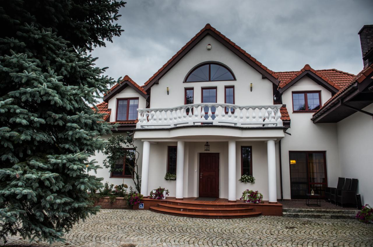 Dwupiętrowy budynek z białymi ścianami, czerwonym dachem i kolumnami podtrzymującymi balkon, widoczne wejście z rampą dla niepełnosprawnych oraz brukowany podjazd.