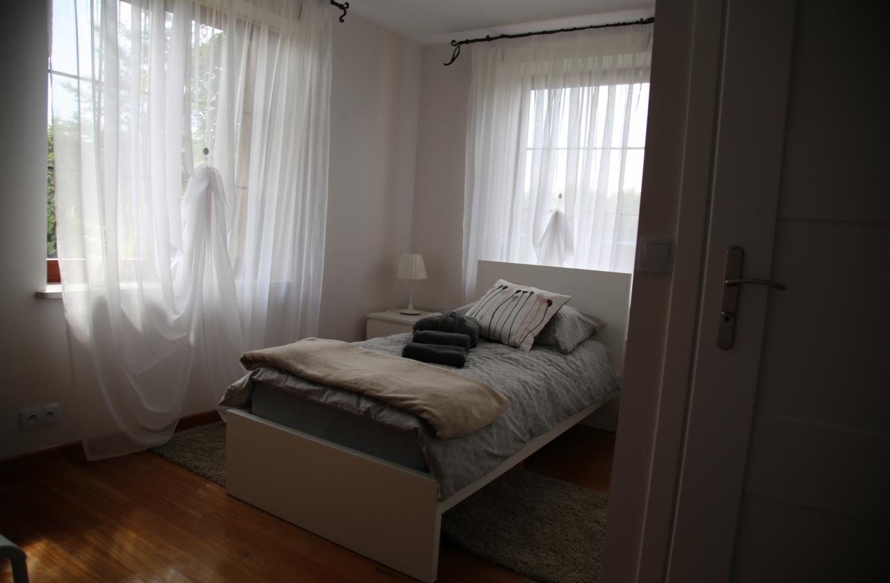 Pokój z łóżkiem jednoosobowym w minimalistycznym stylu, z białymi zasłonami i drewnianą podłogą, widziany z perspektywy otwartych drzwi.