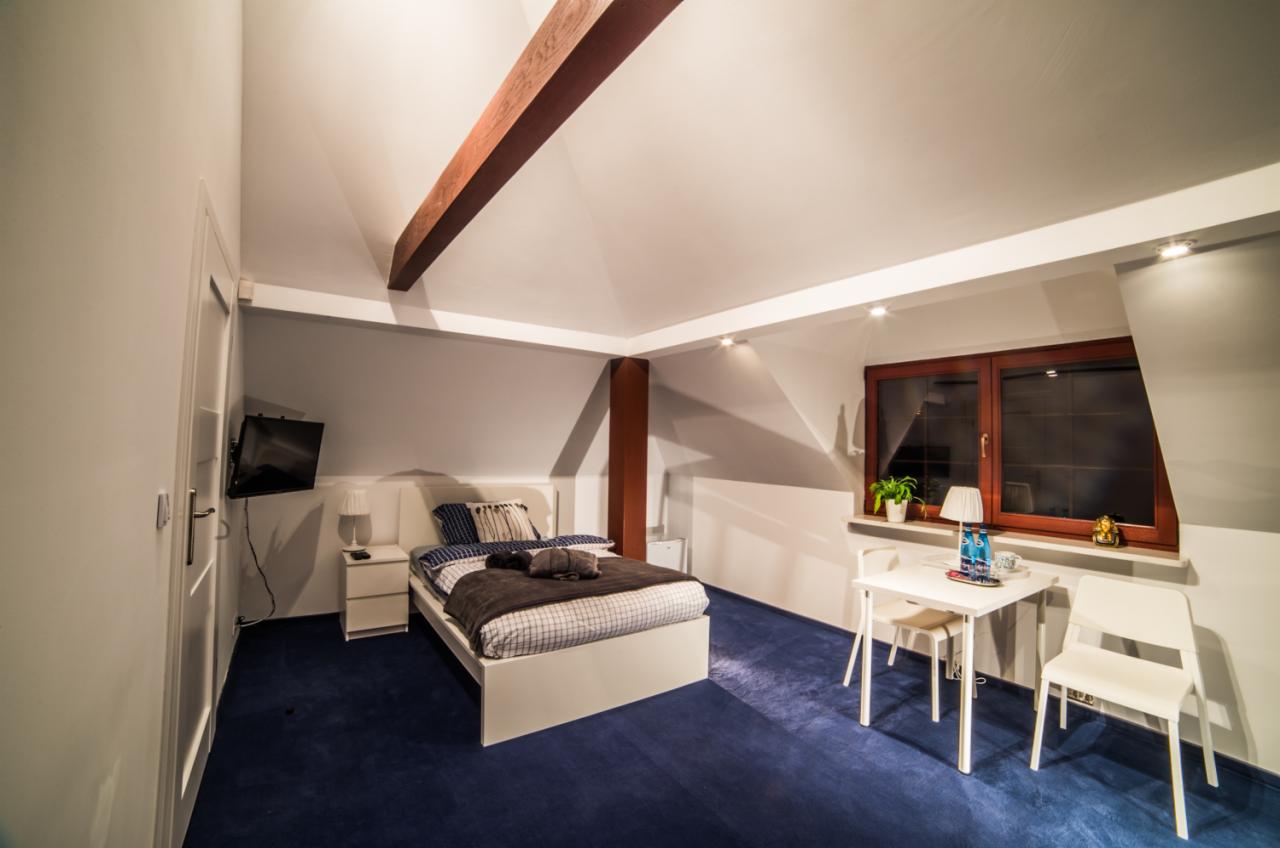 Pokój z łóżkiem, biurkiem i telewizorem, widoczne drewniane belki stropowe i niebieska wykładzina podłogowa.