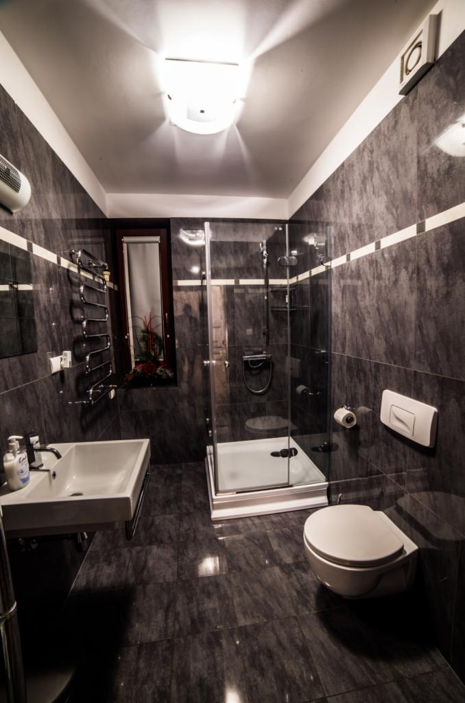 Nowoczesna łazienka z kabiną prysznicową, umywalką i toaletą, wykończona w ciemnych płytkach z jasnymi akcentami, widok na okno z roślinami.