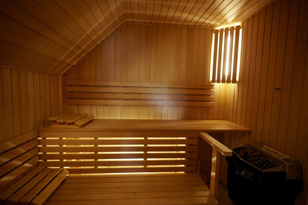Wnętrze sauny wykończone jasnym drewnem, z ławkami na różnych poziomach, oświetleniem za oparciami i piecem do sauny w rogu.
