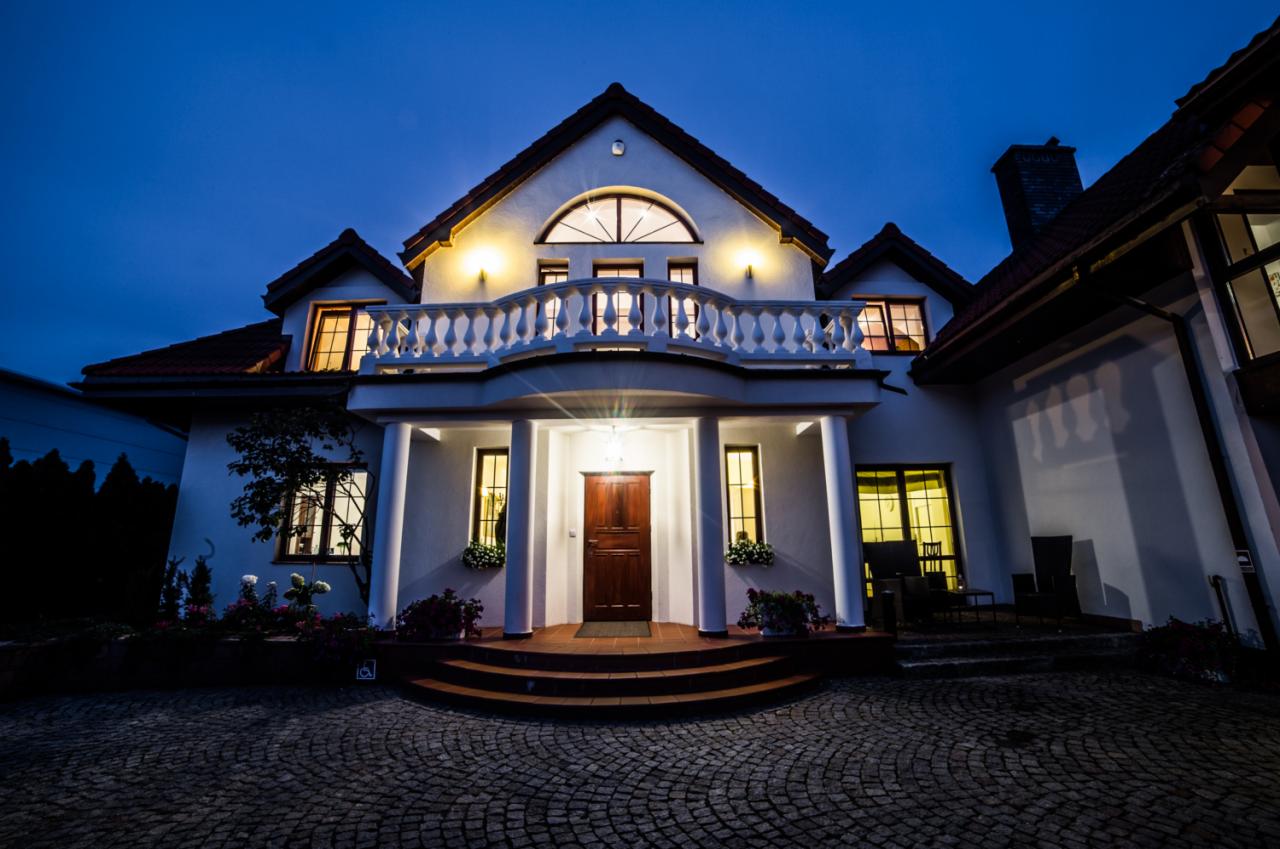 Dwupiętrowy, biały dom z balkonem i oświetlonym wejściem, sfotografowany o zmierzchu, z brukowanym podjazdem.