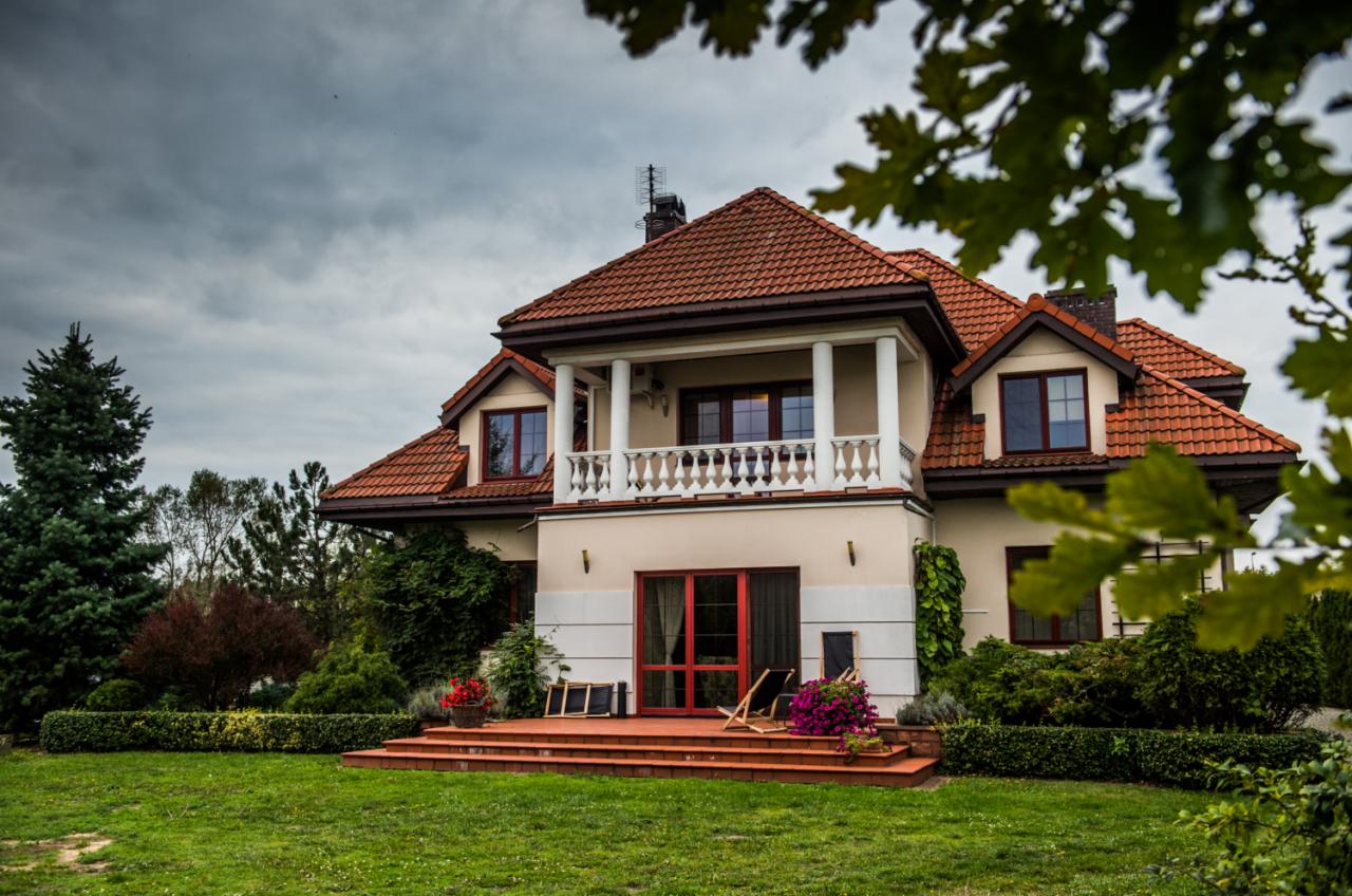 Dwupiętrowy dom z czerwonym dachem i balkonem, otoczony zielonym trawnikiem i roślinnością w pochmurny dzień.