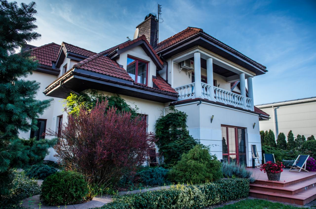 Dwupiętrowy dom z czerwonym dachem i kolumnami na balkonie, otoczony zielenią i ozdobnymi krzewami.
