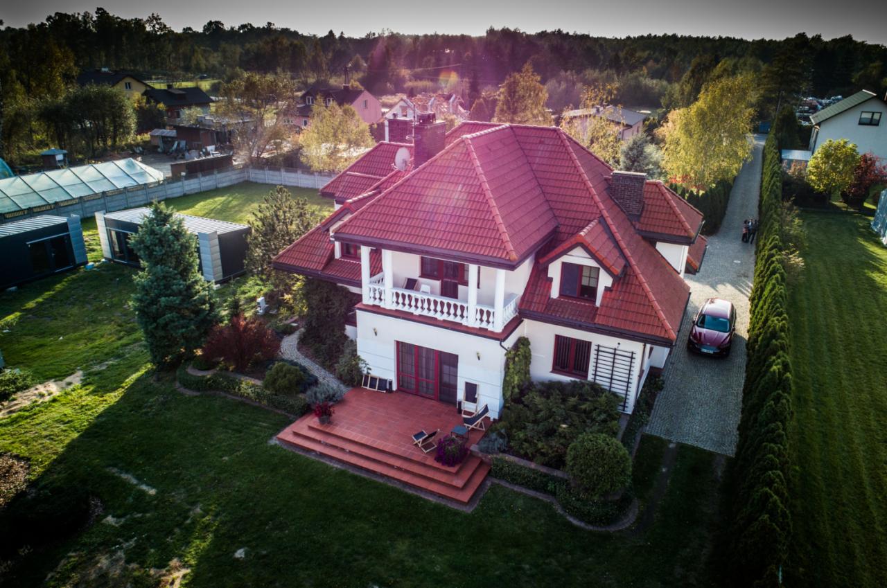 Widok z lotu ptaka na duży, biały dom z czerwoną dachówką, tarasem z czerwonej cegły i zadbanym ogrodem, z samochodem zaparkowanym na podjeździe.