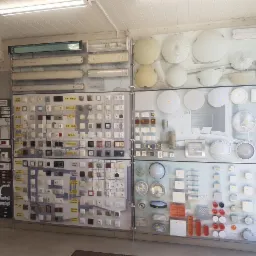 Ekspozycja różnorodnych włączników, gniazdek, lamp sufitowych oraz świetlówek w sklepie elektrycznym, z widocznymi nazwami producentów i modeli na etykietach.