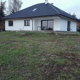 Nowoczesny, parterowy dom z białymi ścianami i ciemną dachówką, widoczny z perspektywy trawnika przed budynkiem, z figurką na kominie.