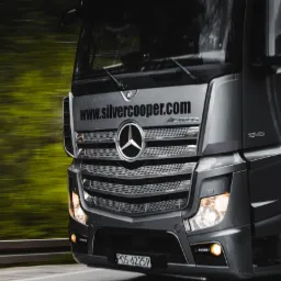 Szary ciągnik siodłowy Mercedes-Benz Actros z widoczną stroną internetową na osłonie chłodnicy, sfotografowany w Pajęcznie podczas jazdy, z rozmytym zielonym tłem.