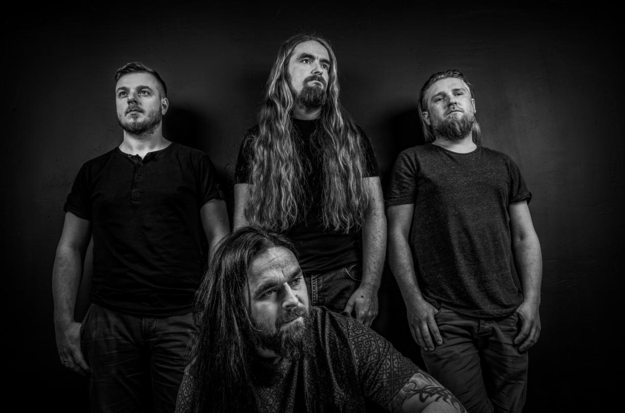 Czarno-białe portretowe zdjęcie czterech mężczyzn z brodami i długimi włosami, ubranych w ciemne koszulki na tle ciemnej ściany, z różnymi wyrazami twarzy.