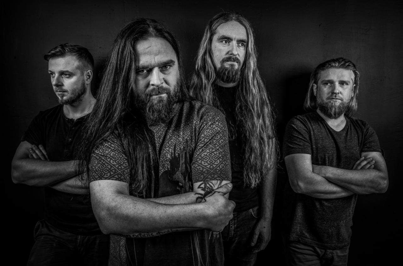 Czarno-białe portretowe zdjęcie czterech mężczyzn z brodami i długimi włosami, stojących na ciemnym tle, z których jeden ma tatuaż na ramieniu i wzorzystą koszulę.