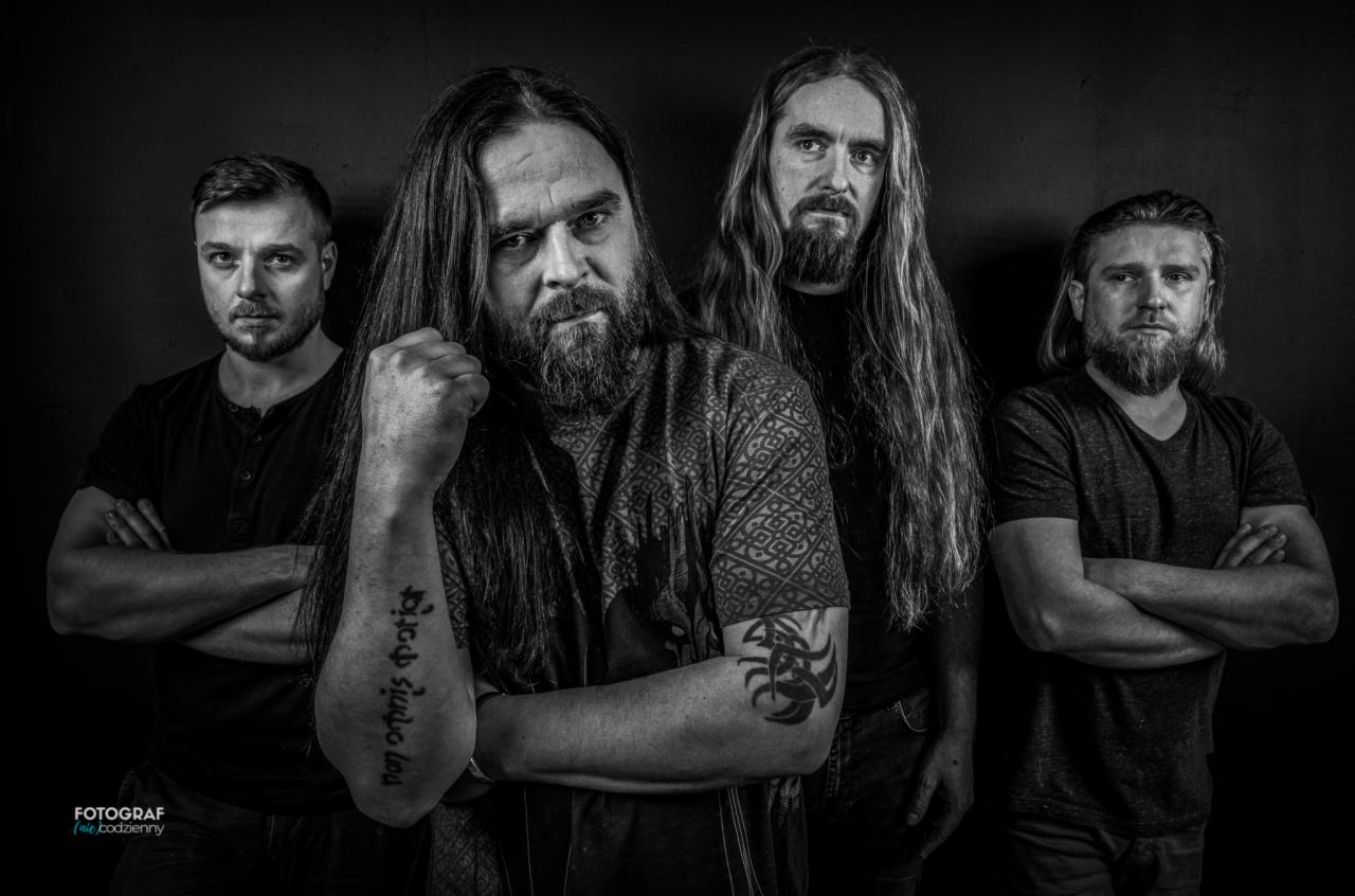 Czarno-białe zdjęcie czterech mężczyzn z brodami i długimi włosami, ubranych w ciemne ubrania, stojących na ciemnym tle. Jeden z mężczyzn ma wytatuowane ramię z napisem w obcym języku i symbolem.