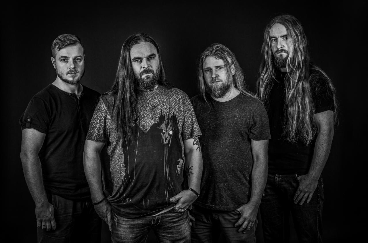 Czarno-białe portretowe zdjęcie studyjne czterech mężczyzn z długimi włosami i brodami, ubranych w codzienne ubrania, na ciemnym tle.