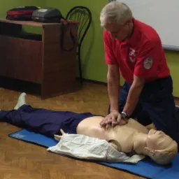 Instruktor w czerwonej koszulce z logo Czerwonego Krzyża demonstruje resuscytację krążeniowo-oddechową na manekinie podczas kursu pierwszej pomocy. Na stole w tle widoczne wyposażenie medyczne.