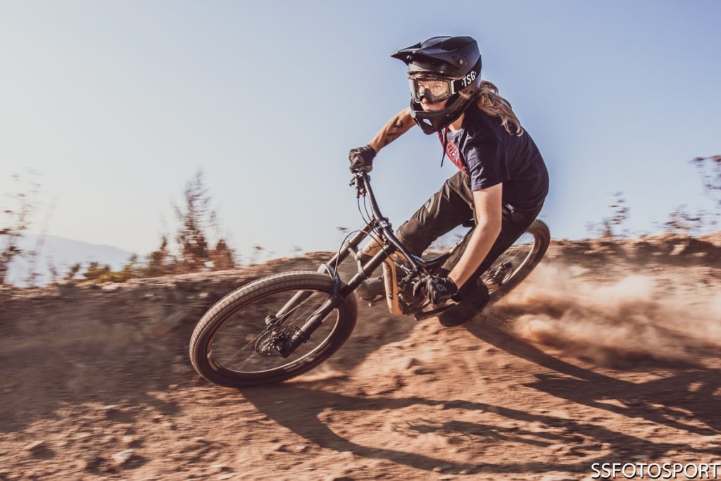 Kobieta w kasku i goglach rowerowych dynamicznie pokonuje trasę na rowerze górskim, unosząc tumany kurzu.