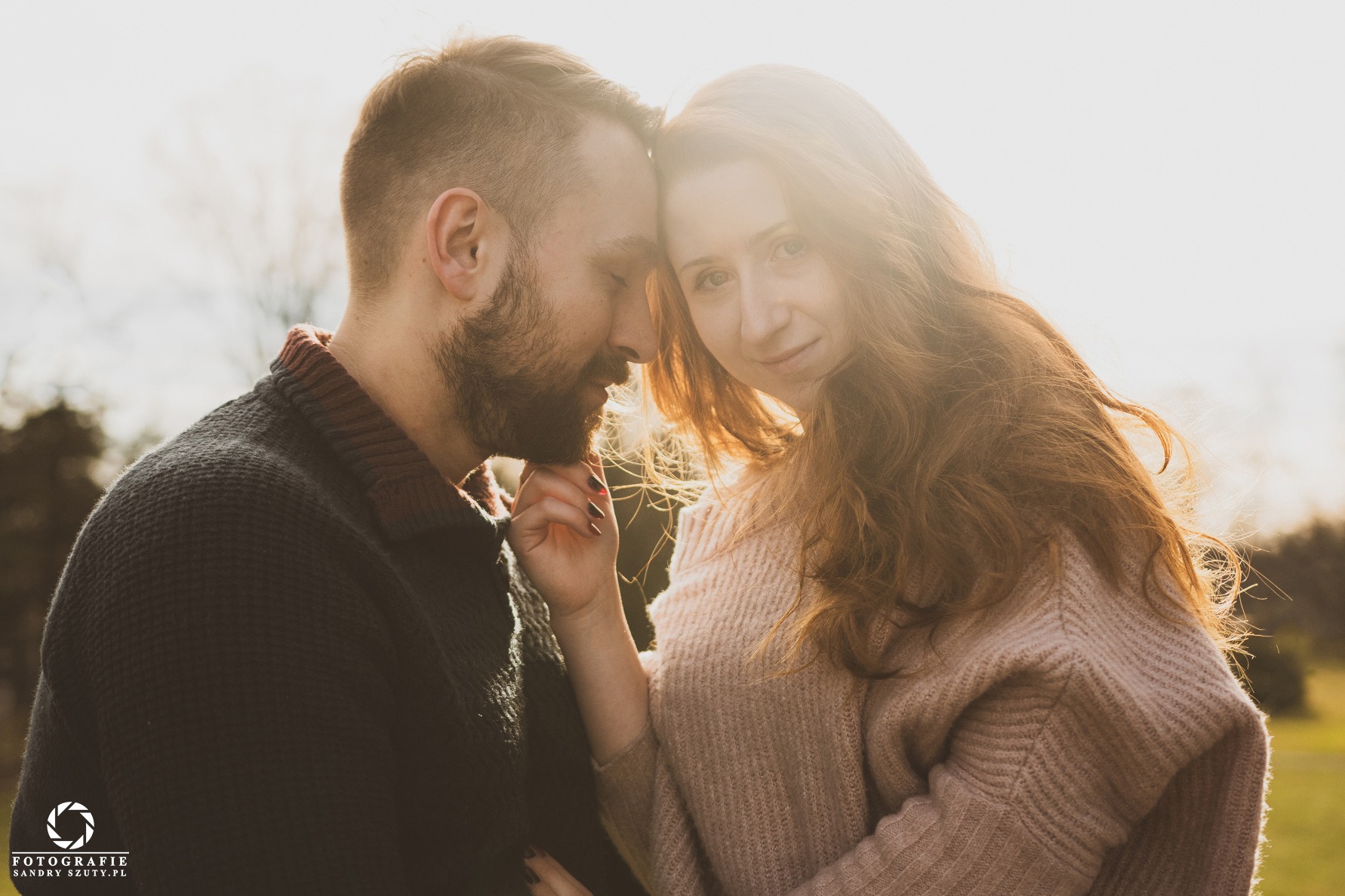 Zakochana para, mężczyzna z brodą i kobieta z rudymi włosami, stykają się czołami w ciepłym świetle słonecznym, ona patrzy w obiektyw.