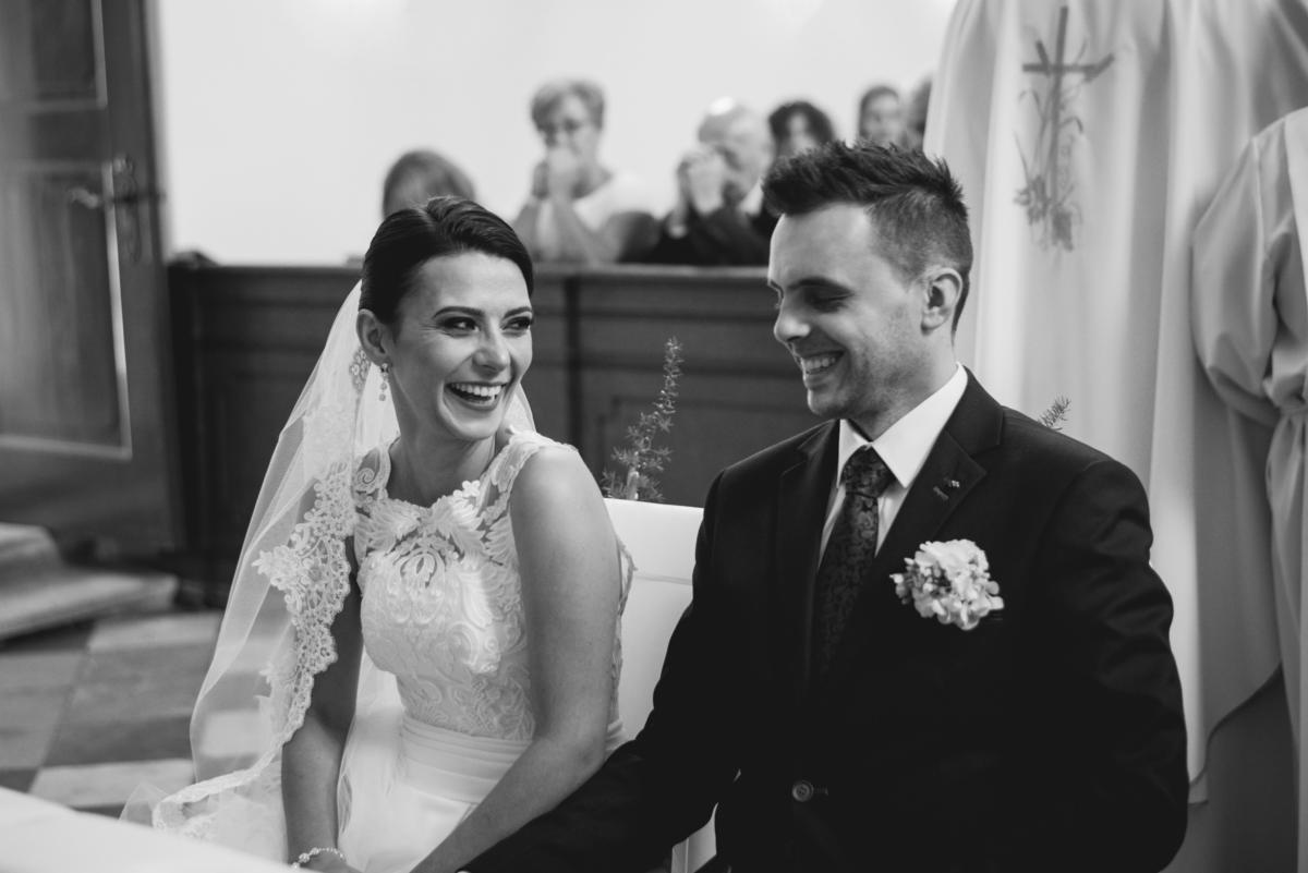 Czarno-białe zdjęcie uśmiechniętej panny młodej w koronkowej sukni i welonie siedzącej obok uśmiechniętego pana młodego w ciemnym garniturze z butonierką podczas ceremonii ślubnej w kościele.