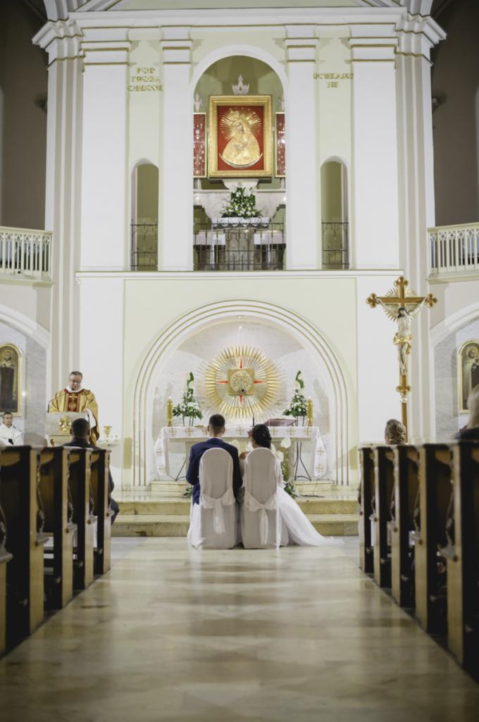 Para młoda siedzi przed ołtarzem w kościele podczas ceremonii ślubnej, widok z tyłu, z dekorowanymi krzesłami i długim welonem panny młodej, ksiądz odprawia mszę w tle.