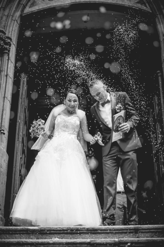 Czarno-białe zdjęcie nowożeńców wychodzących z kościoła, obsypywanych confetti. Panna młoda w sukni z koronkową górą i bukietem. Pan młody w smokingu z muszką, trzyma obrazek religijny.