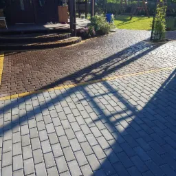 Ułożona ścieżka z kostki brukowej w dwóch kolorach: szarym i brązowym, oddzielona żółtą linią, prowadząca do domu z drewnianym tarasem, cień drzewa na kostce.