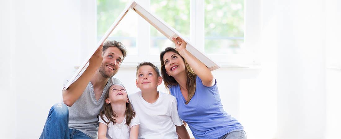 Szczęśliwa rodzina z dwójką dzieci siedzi na podłodze w jasnym pokoju, trzymając nad głowami ramę obrazu, tworząc symboliczny dach domu i patrząc w górę z uśmiechem.