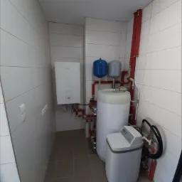 Instalacja kotła grzewczego z zbiornikiem akumulacyjnym, naczyniami wzbiorczymi i zmiękczaczem wody w kotłowni wyłożonej białymi kafelkami.