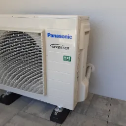 Zewnętrzna jednostka klimatyzacji Panasonic Inverter z widocznym logo i oznaczeniem R32, zamontowana na czarnych podkładkach antywibracyjnych na szarej kostce brukowej przy jasnej ścianie.