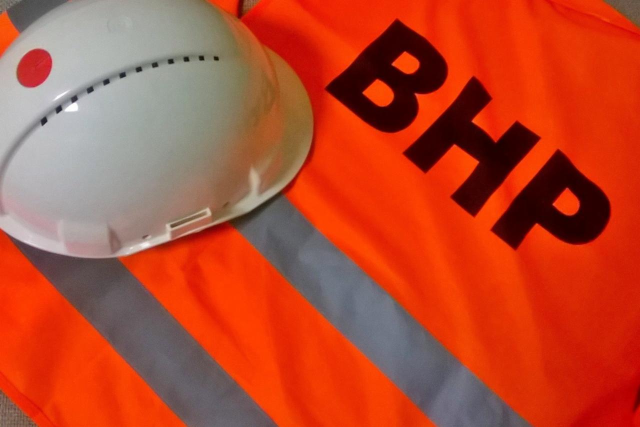 Biały kask ochronny z czerwoną naklejką i czarnymi otworami wentylacyjnymi leży na pomarańczowej kamizelce odblaskowej z napisem BHP.