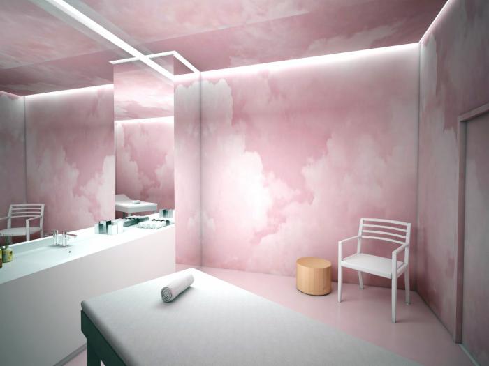 Jasne wnętrze gabinetu kosmetycznego z łóżkiem do masażu, białym krzesłem i ścianami w pastelowym różu z motywem chmur, odbicie w lustrze kolejnego łóżka.