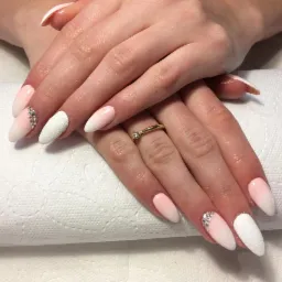 Dłonie z delikatnym, ombre manicure w odcieniach różu i bieli, jeden paznokieć pokryty białą, teksturowaną powłoką, a drugi zdobiony drobnymi kryształkami, na palcu złoty pierścionek.