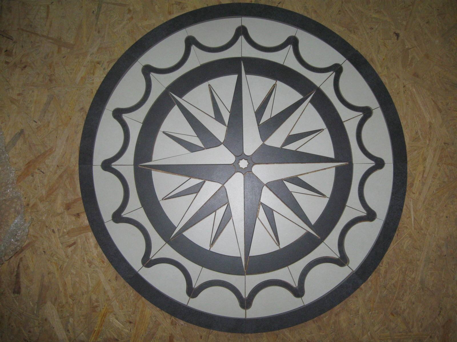 Okrągła mozaika podłogowa z motywem kompasu w odcieniach szarości i bieli, ułożona na płycie OSB. Widoczne fugi i detale wzoru.