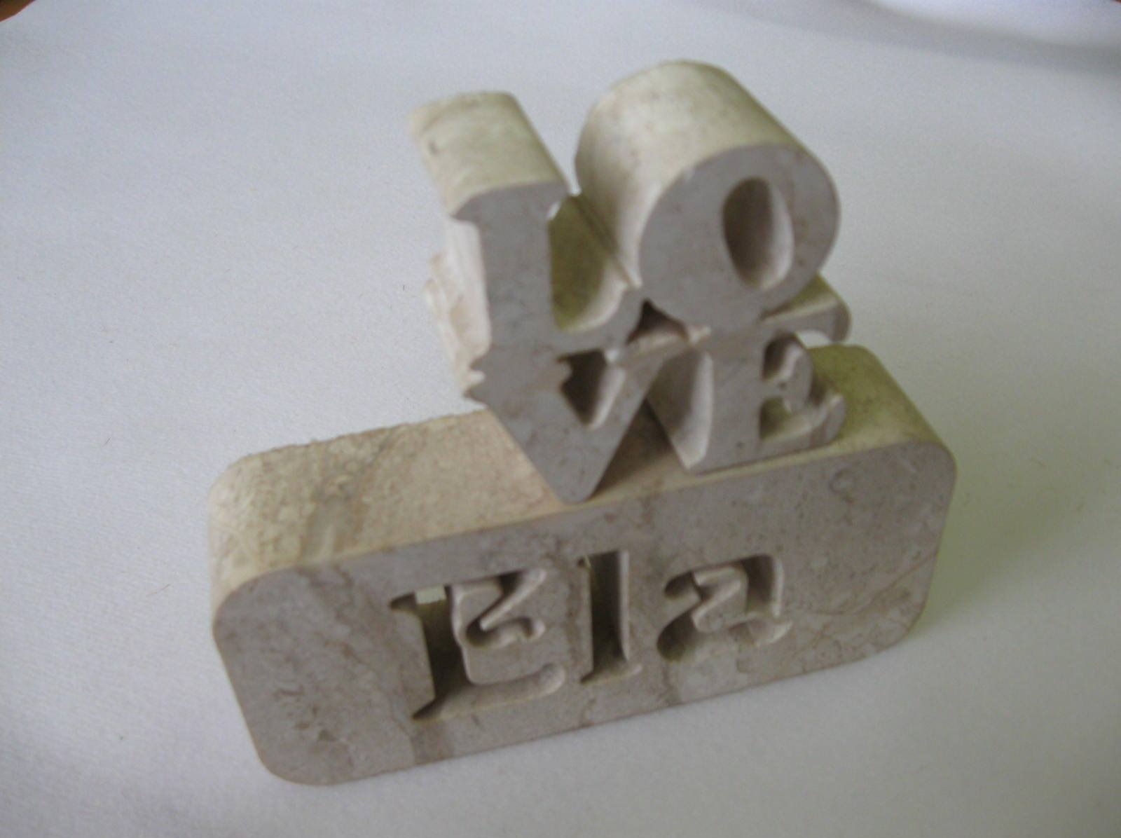 Ręcznie wykonana, kamienna rzeźba z napisem 'LOVE' umieszczona na podstawie z wyciętym imieniem 'Ela'. Jasne tło, delikatne światło.