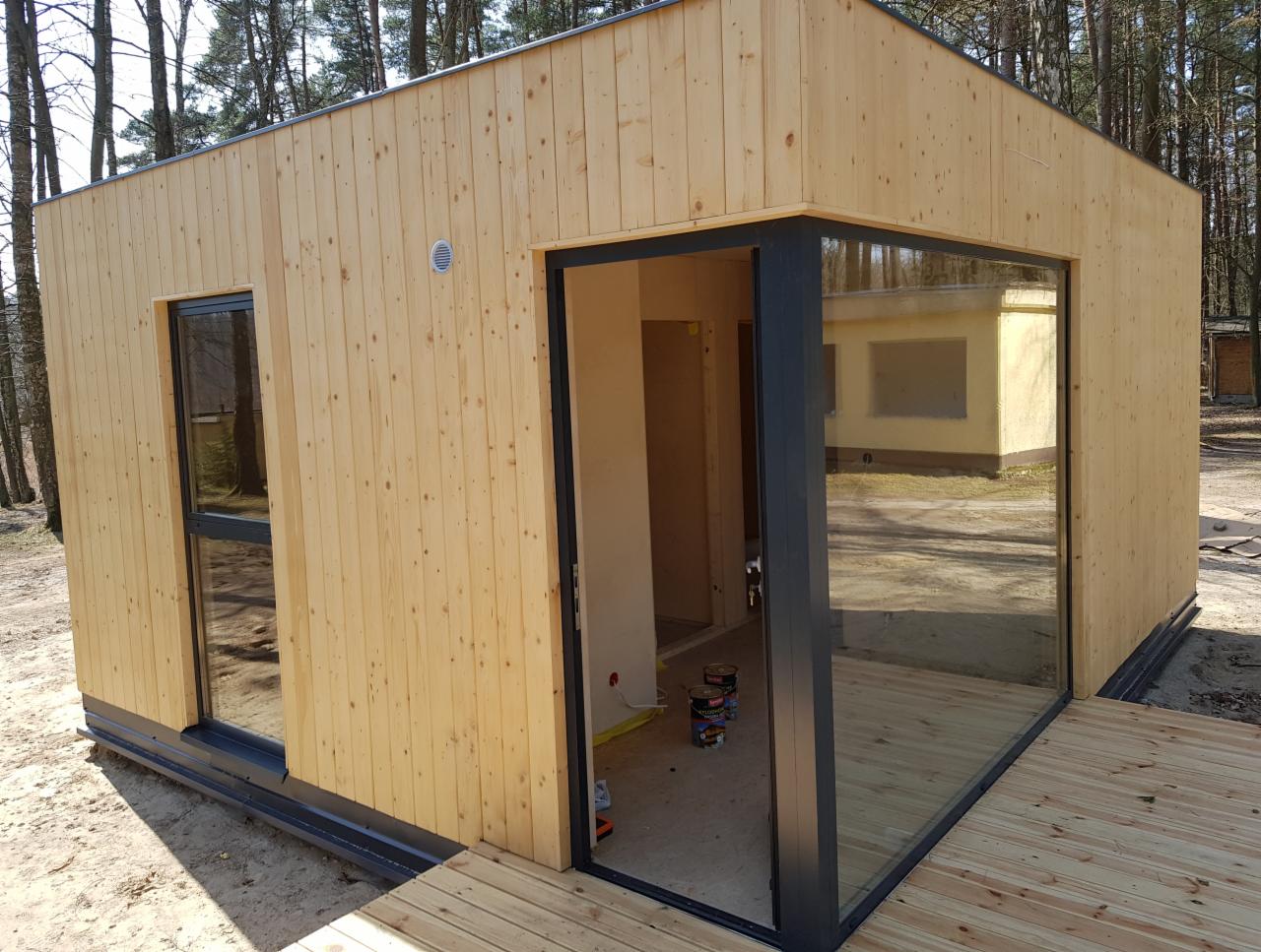 Surowy, drewniany budynek z dużym narożnym oknem, widoczne puszki farby wewnątrz, otoczenie leśne, częściowo wykończony taras.