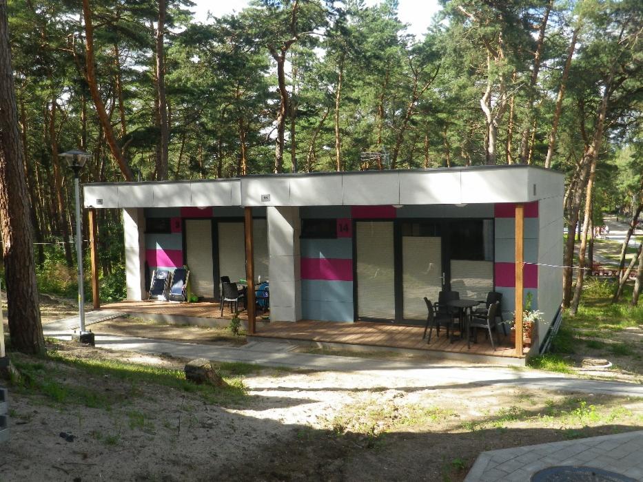 Nowoczesny, parterowy budynek rekreacyjny z drewnianym tarasem i kolorowymi akcentami na elewacji, otoczony sosnowym lasem.