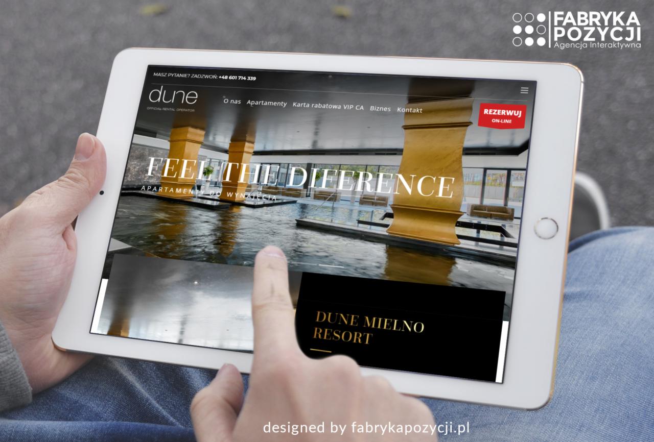 Tablet wyświetlający stronę internetową apartamentów Dune Mielno Resort, z palcem wskazującym na ekranie, w tle logo agencji interaktywnej Fabryka Pozycji.