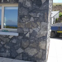 Ściana budynku wykończona nieregularnymi kamieniami w odcieniach szarości i czerni, widoczne okno z odbijającym się niebem i morzem, obok zaparkowana ciężarówka z gruzem.